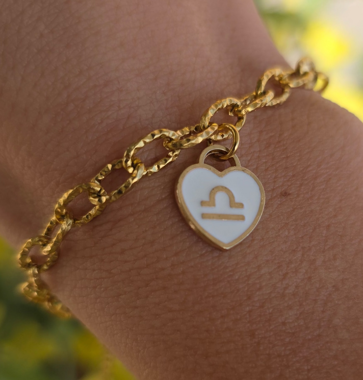 Bracelet doré avec pendentif cœur blanc représentant un signe astrologique, porté au poignet, en gros plan.